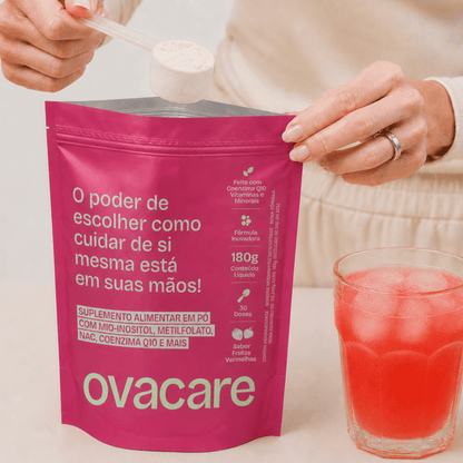 Fotografia estilo lifestyle mostrando o preparo do suplemento Ovacare SOMP. Uma mulher segura um dosador transparente com o pó do suplemento sobre a embalagem cor-de-rosa aberta. Ao lado, sobre uma mesa clara, há um copo de vidro com a bebida já misturada, de cor vermelha vibrante e com pedras de gelo.